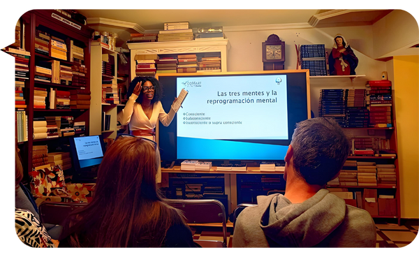 Niurys Núñez dando una conferencia sobre reprogramación mental en una sala con estanterías llenas de libros, con una presentación proyectada en una pantalla y un público atento.