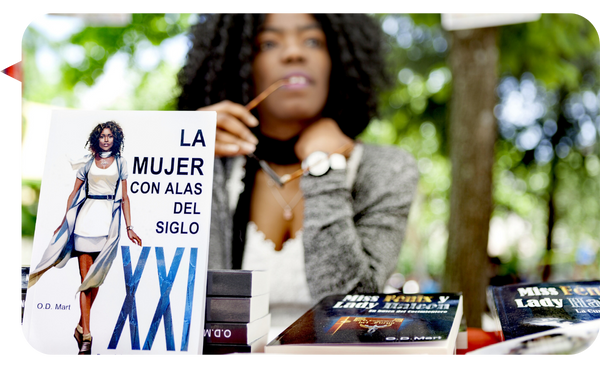 Niurys Núñez en un evento literario al aire libre, rodeada de libros, con su obra "La Mujer con Alas del Siglo XXI" en primer plano, mientras sostiene un bolígrafo con una expresión reflexiva.