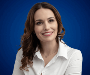 Fotografía de Eva Mazuecos con camisa blanca y fondo azul, sonriendo con los brazos cruzados en una pose profesional.