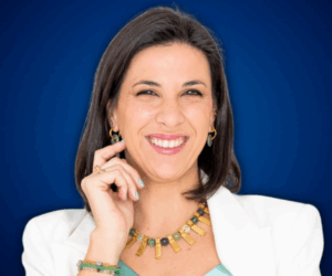 Fotografía de Gemma Ramírez con fondo azul profesional, luciendo una chaqueta blanca, collar dorado y una sonrisa segura y carismática.