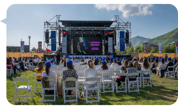 Audiencia al aire libre en un evento con un escenario grande y pantallas, con montañas de fondo.