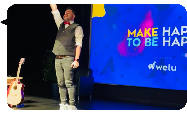 Juan Carlos Gutiérrez en el escenario durante una conferencia motivacional sobre felicidad, levantando la mano con una guitarra al fondo.